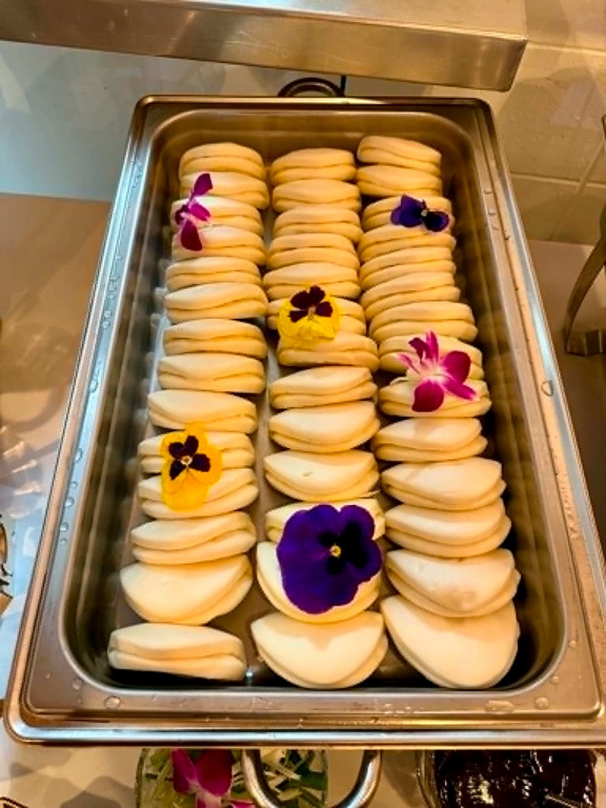 Steamed bao buns in a metal chafing tray, garnished with edible pansy flowers