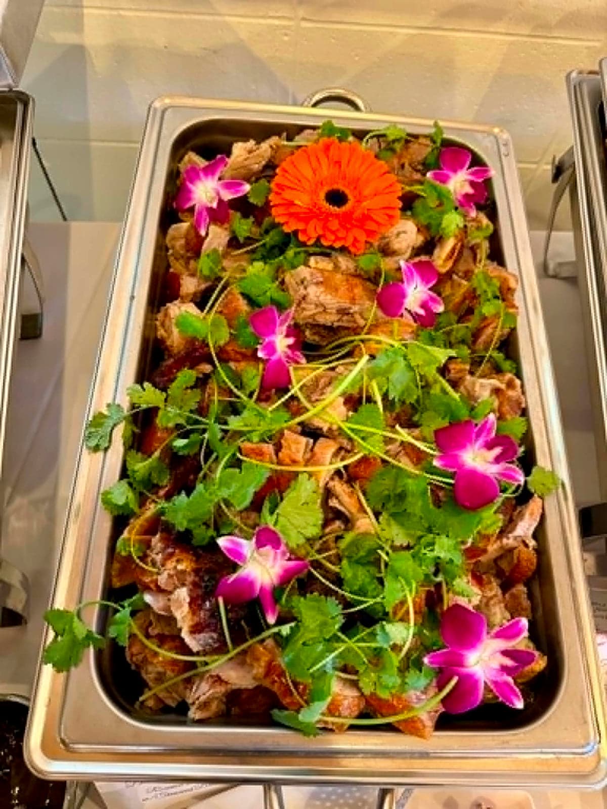 Tray of roasted meat topped with cilantro, orchids and an orange gerbera flower